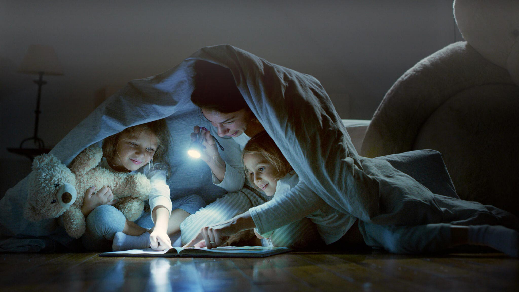 Mutter sitzt mit zwei Kindern im Dunkeln unter einer Decke, sie hält eine Taschenlampe in der Hand.