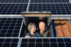 Isabelle und Lukas Loosli blicken aus dem Dachfenster, rund um sie sieht man die Panels der Solaranlage.