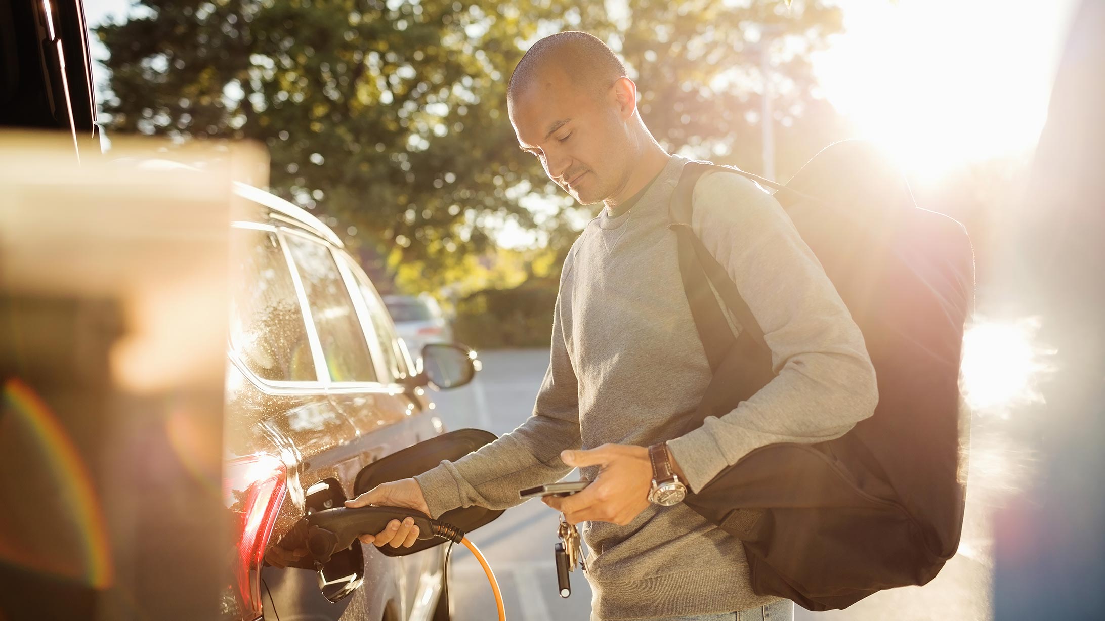 Ein Mann mit Sporttasche blickt auf sein Handy und zieht dabei den Stecker seiner eigenen Ladestation aus dem Auto.