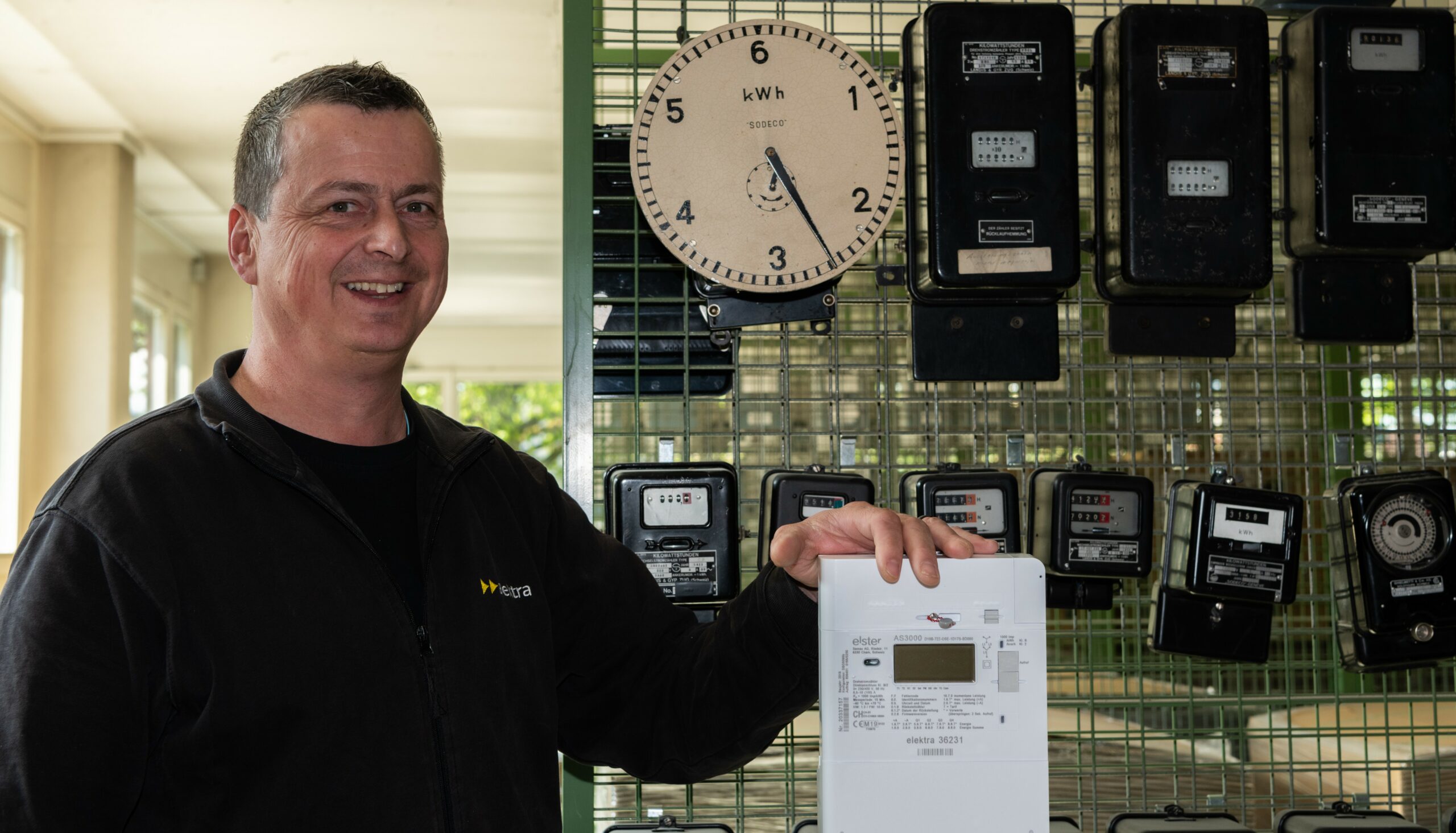 Servicemonteur Bruno Suter steht vor einer Wand voller alter Stromzähler und hält einen modernen Smart Meter in der Hand.